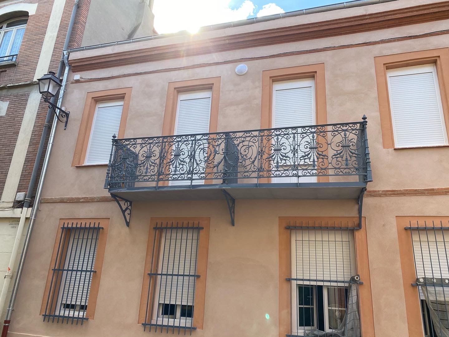 Balcon en fer forgé, fenêtres avec volets, façade en stuc.